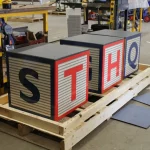 Three large cube blocks with the letters S, T, H, and Q on visible sides are placed on a wooden pallet in an industrial workshop setting.