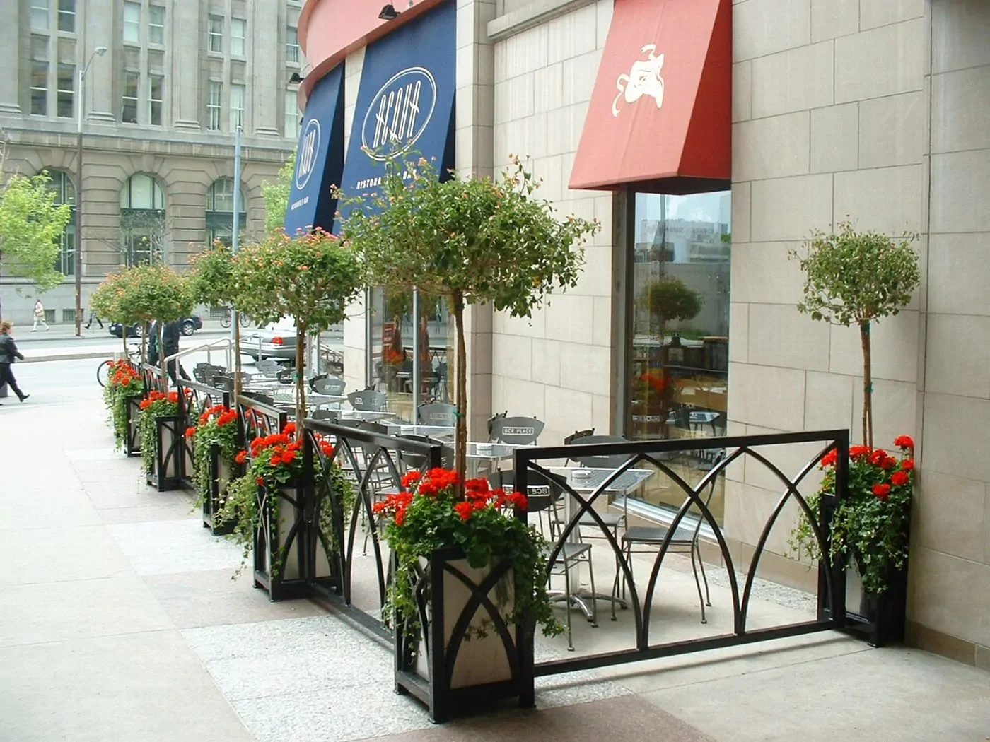 Outdoor café seating area with metal tables and chairs, surrounded by planters with small trees and red flowers, located on a city sidewalk next to a building.