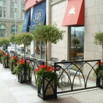 Outdoor café seating area with metal tables and chairs, surrounded by planters with small trees and red flowers, located on a city sidewalk next to a building.