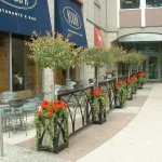 Outdoor restaurant seating area with metal tables and chairs, separated from the walkway by planters holding small trees and red flowers, next to a building entrance.