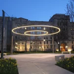 A circular light installation is suspended above a paved plaza with benches and flowerbeds in front of a large stone building at dusk.