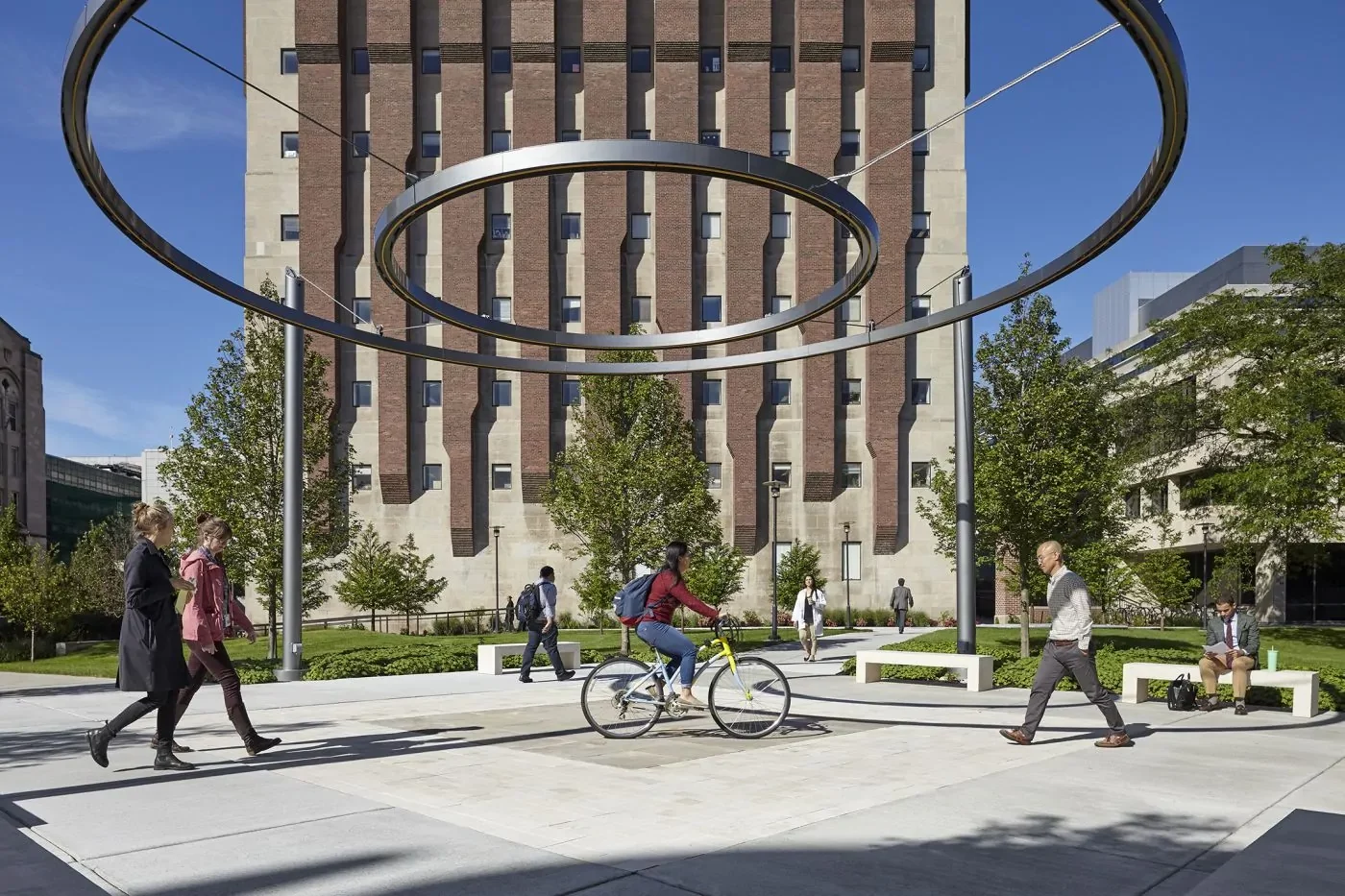 People walk and cycle through a modern outdoor plaza with large metal rings overhead, surrounded by trees and a tall brick building in the background.