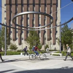 People walk and cycle through a modern outdoor plaza with large metal rings overhead, surrounded by trees and a tall brick building in the background.