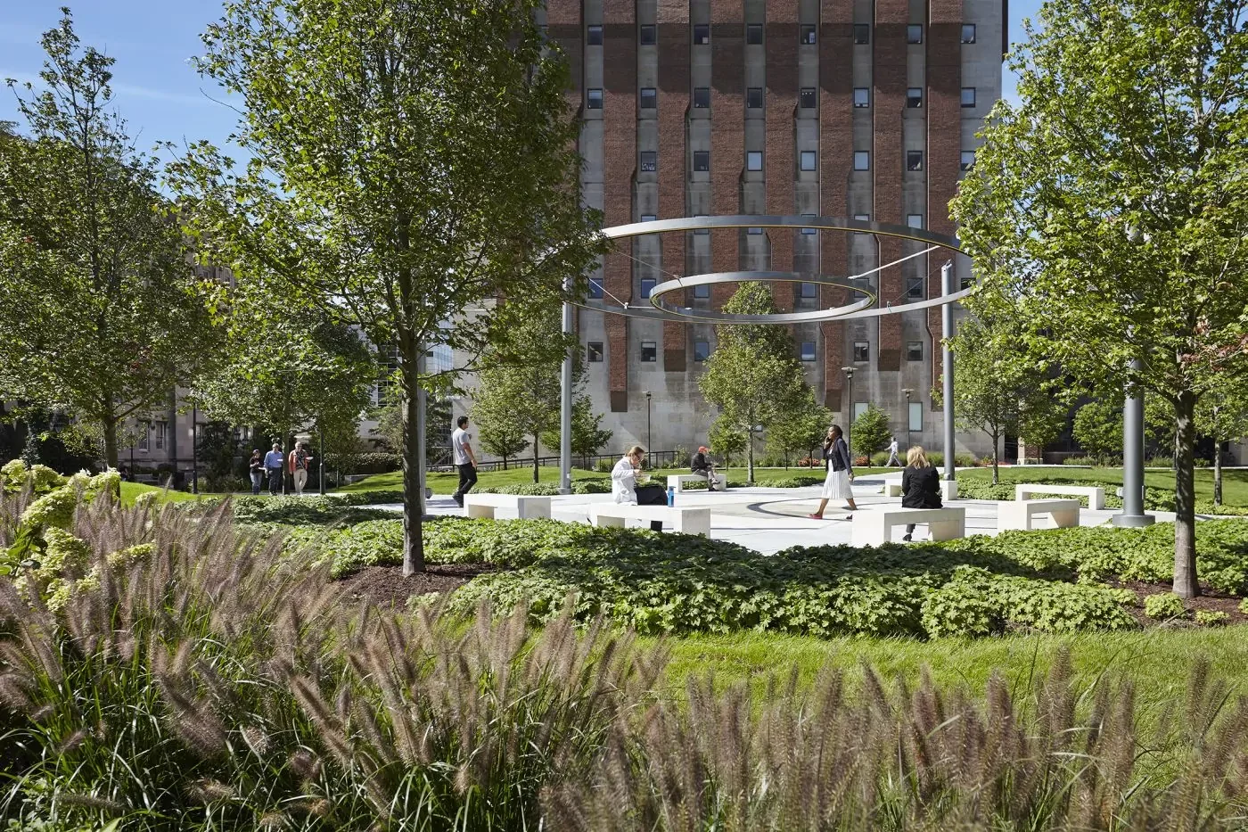 People sit and walk around a circular modern sculpture in a landscaped garden with trees and greenery, in front of a tall brick building.