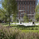 People sit and walk around a circular modern sculpture in a landscaped garden with trees and greenery, in front of a tall brick building.