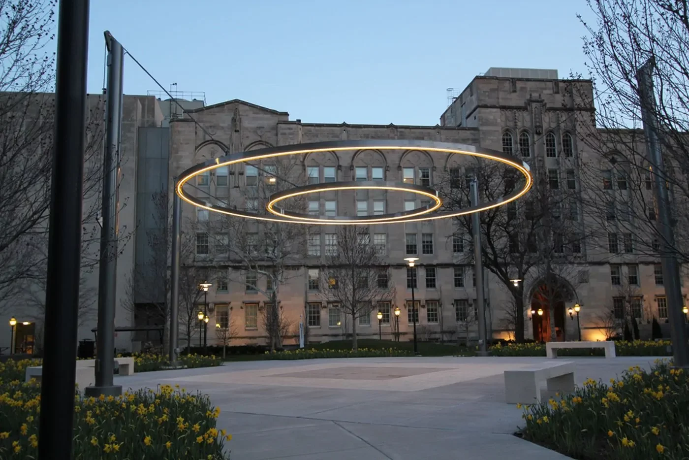 A modern art installation with two circular LED light rings is suspended above a courtyard in front of a stone building at dusk.