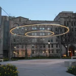 A modern art installation with two circular LED light rings is suspended above a courtyard in front of a stone building at dusk.