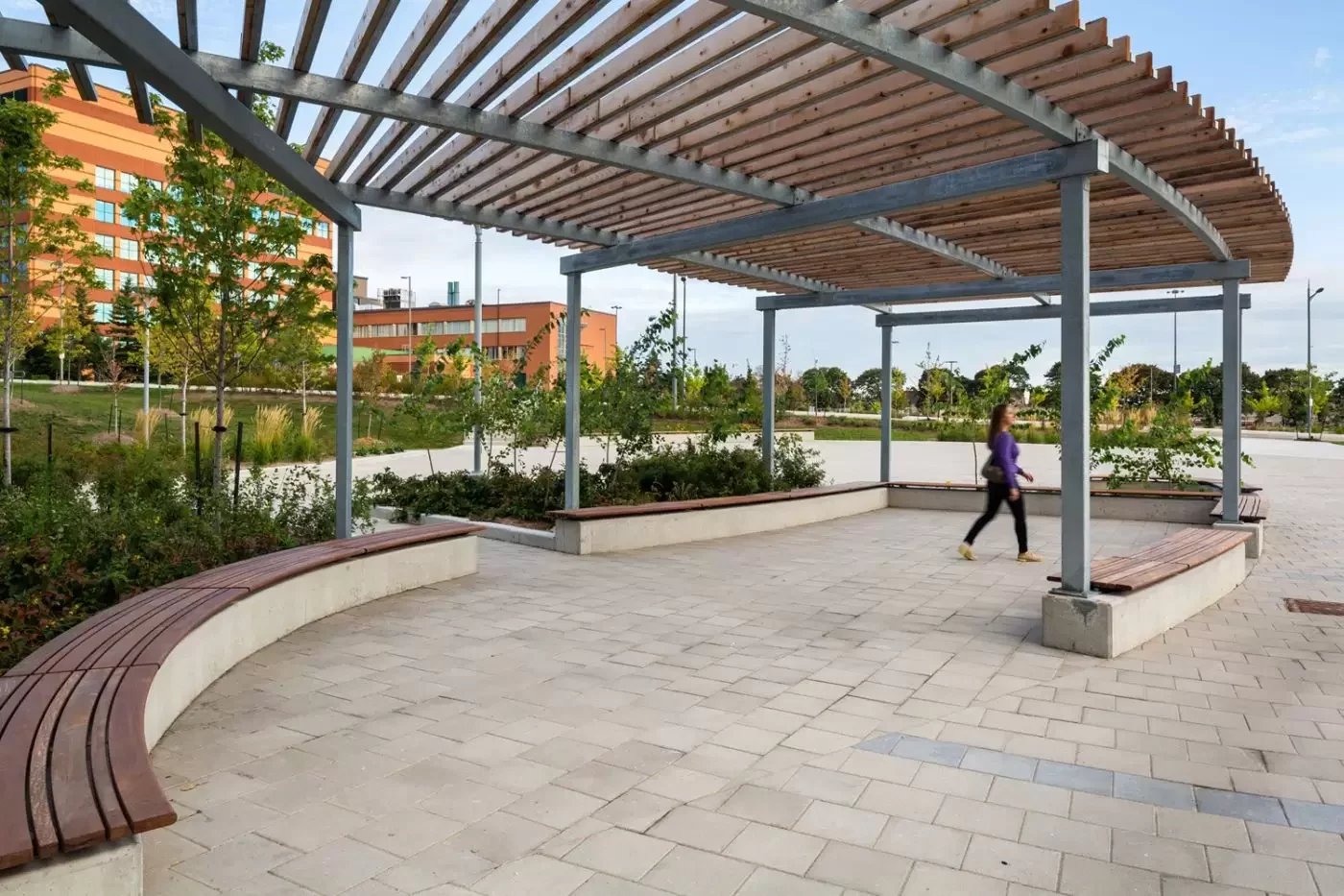 A person walks under a large pergola with wooden slats and curved benches in a landscaped outdoor plaza near modern buildings.