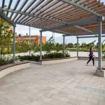A person walks under a large pergola with wooden slats and curved benches in a landscaped outdoor plaza near modern buildings.