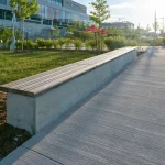 A long, concrete and wood bench lines a paved pathway in an urban park with grass, trees, and a modern building in the background.