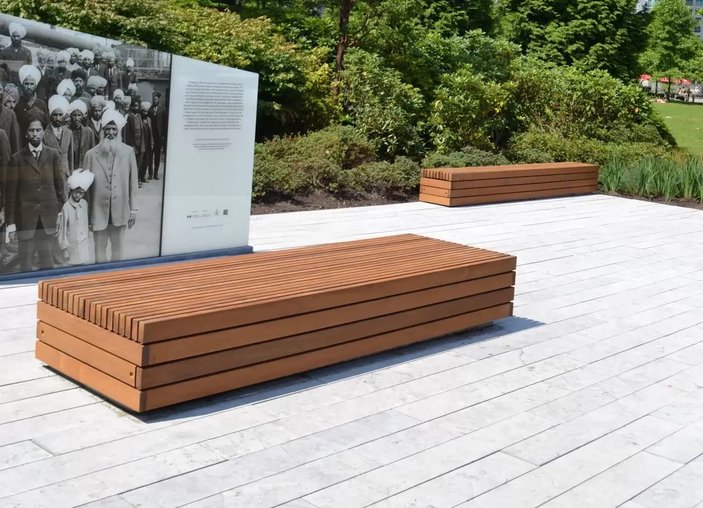 A wooden bench sits on a light-colored paved area next to an informational display with a historical photo; shrubs and trees are in the background.