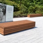 A wooden bench sits on a light-colored paved area next to an informational display with a historical photo; shrubs and trees are in the background.