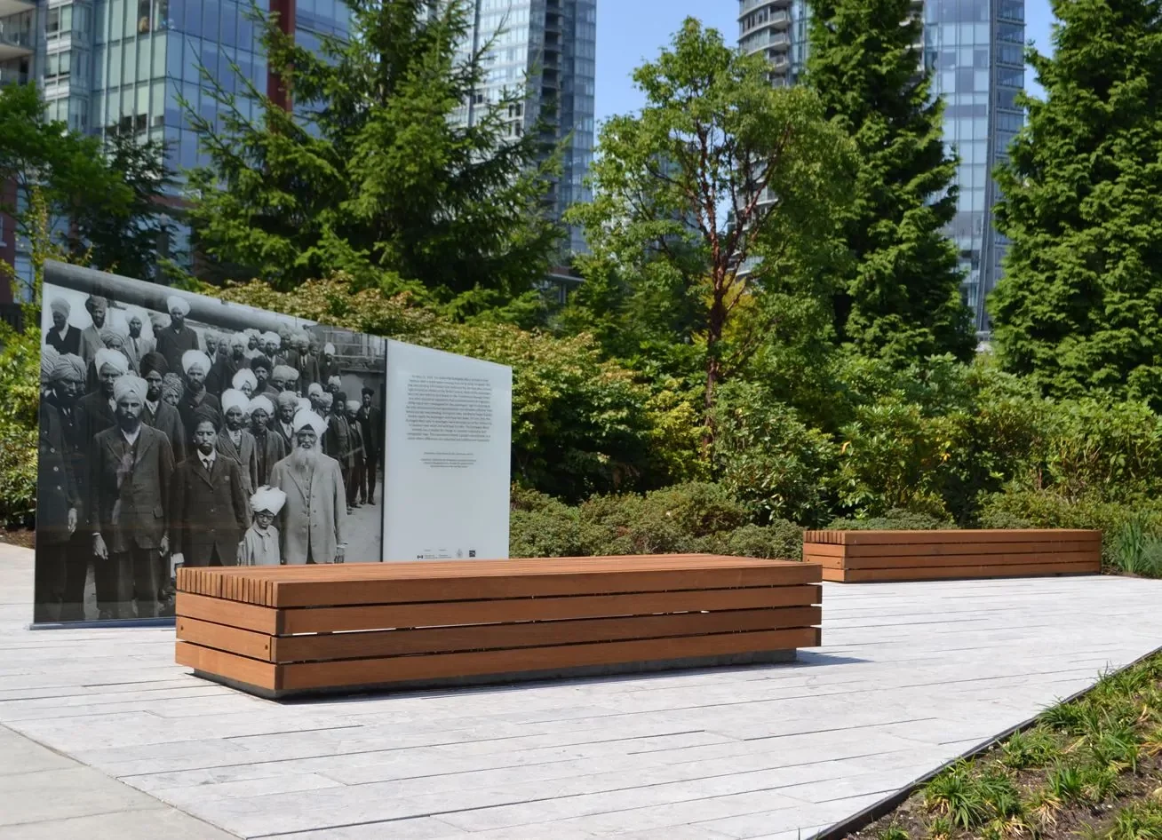 Outdoor memorial with a historical photo display and two wooden benches, surrounded by greenery and tall modern buildings in the background.