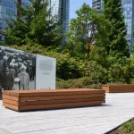 Outdoor memorial with a historical photo display and two wooden benches, surrounded by greenery and tall modern buildings in the background.