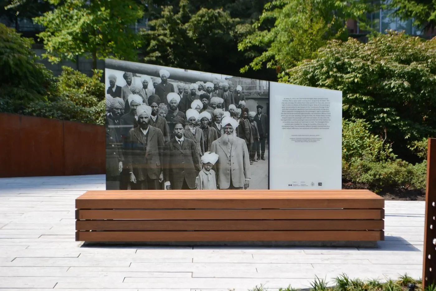 A public outdoor memorial featuring a large historic photograph of a group of men and boys with turbans, displayed on a glass panel beside an explanatory text.