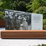 A public outdoor memorial featuring a large historic photograph of a group of men and boys with turbans, displayed on a glass panel beside an explanatory text.