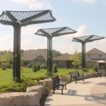 Three metal shade structures with geometric tops stand along a curved sidewalk with benches and stone edging in a grassy park near residential houses.
