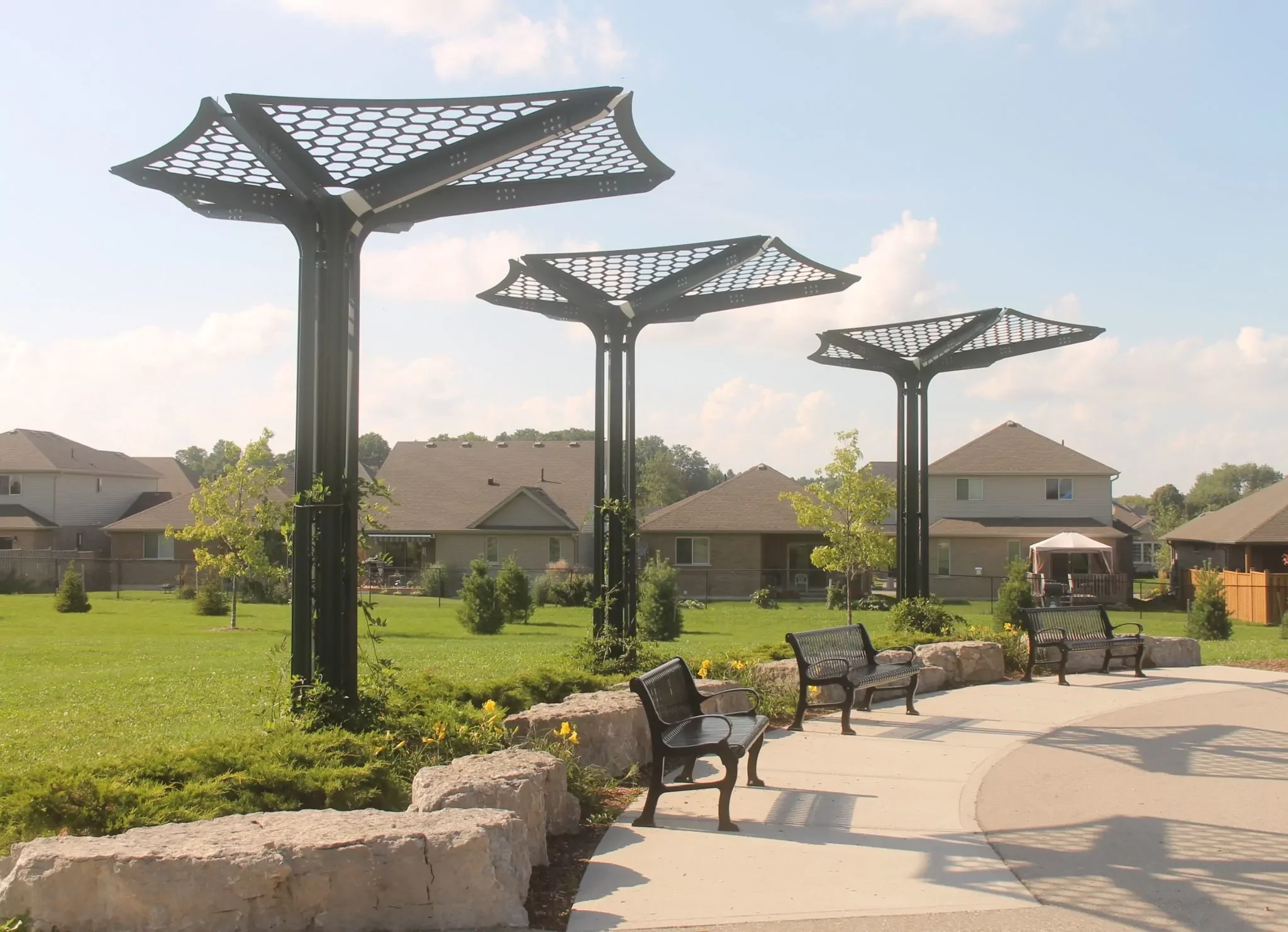 Three metal shade structures with geometric tops stand along a curved sidewalk with benches and stone edging in a grassy park near residential houses.