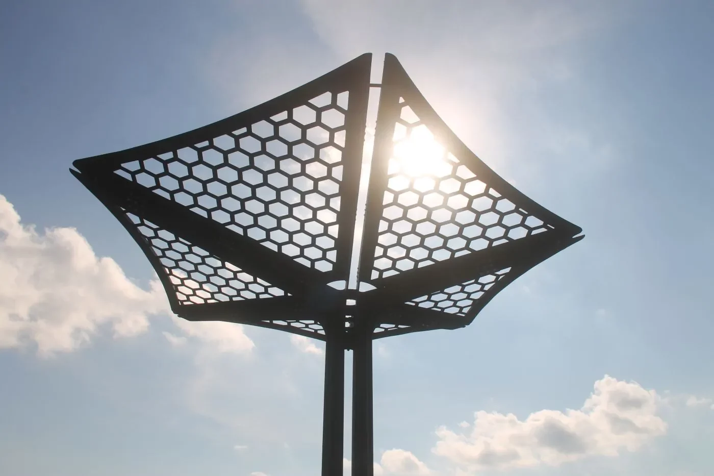 CUSTOM-Living-Shade-Infrastructure-02 Hexagonal-patterned metal structure with a canopy shape is shown against a blue sky with the sun shining through it.