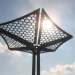 Hexagonal-patterned metal structure with a canopy shape is shown against a blue sky with the sun shining through it.
