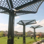 Three metal structures with hexagonal-patterned roofs stand in a grassy park area near suburban houses under a partly cloudy sky.
