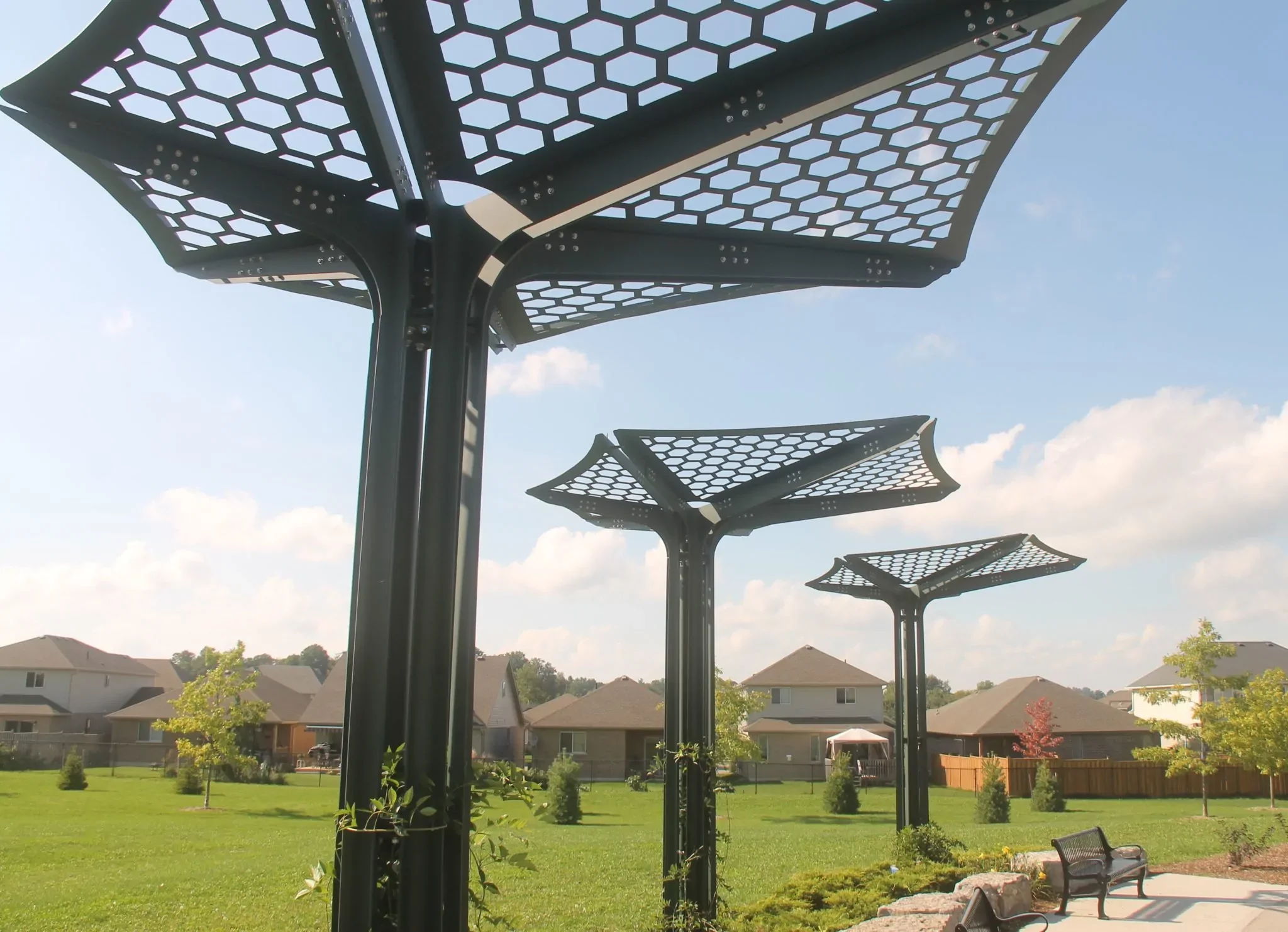 Three metal structures with hexagonal-patterned roofs stand in a grassy park area near suburban houses under a partly cloudy sky.