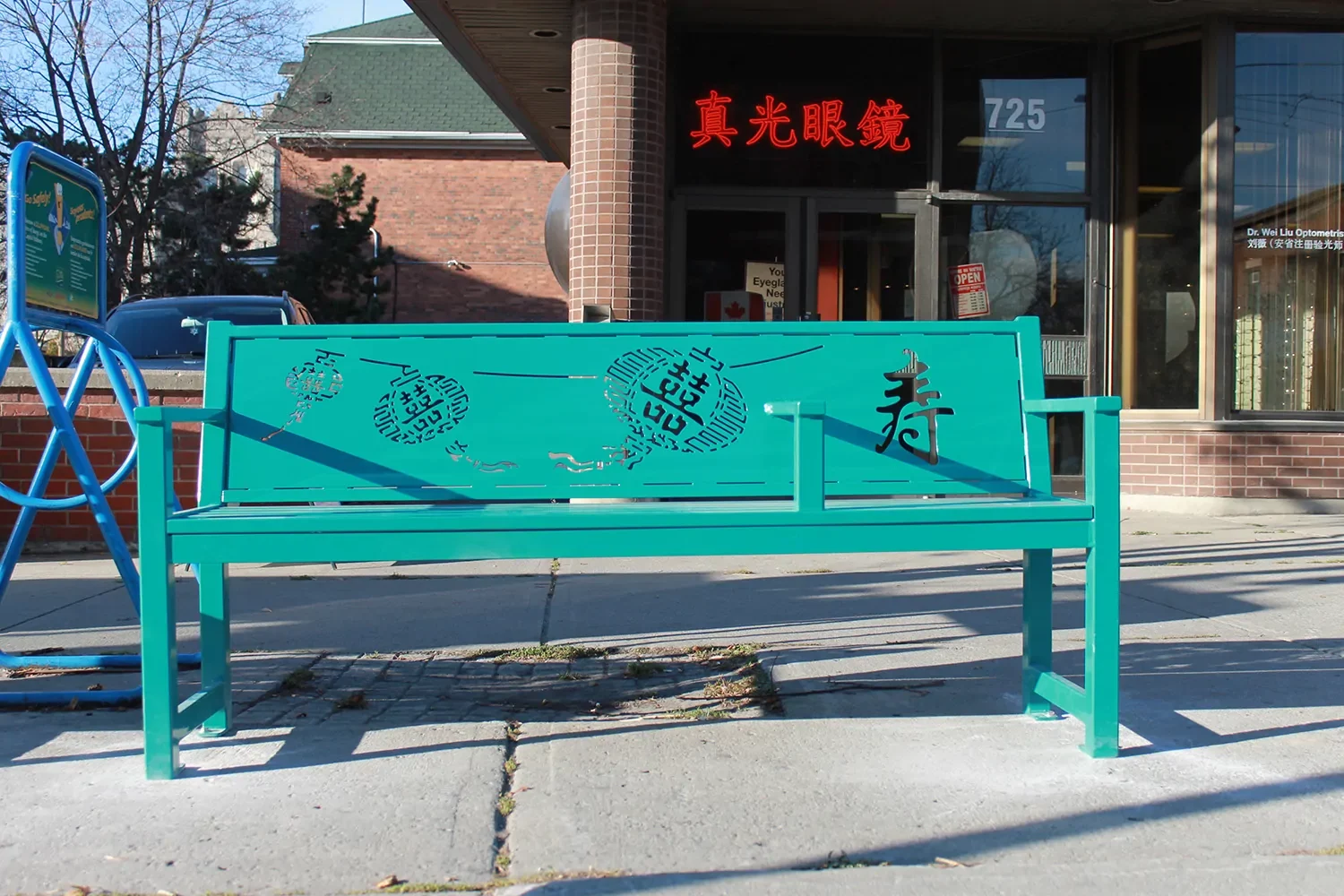 A green bench with decorative Asian calligraphy sits on a sidewalk in front of a brick building with a glass door and red signage in Chinese characters.