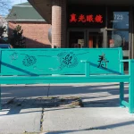 A green bench with decorative Asian calligraphy sits on a sidewalk in front of a brick building with a glass door and red signage in Chinese characters.