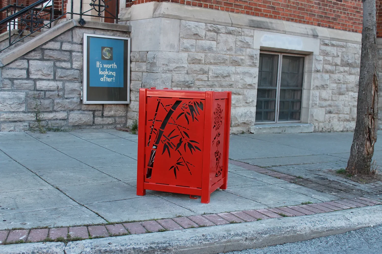 A red metal trash bin with a bamboo design stands on a sidewalk near a brick building and a sign that reads, 