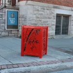 A red metal trash bin with a bamboo design stands on a sidewalk near a brick building and a sign that reads, 