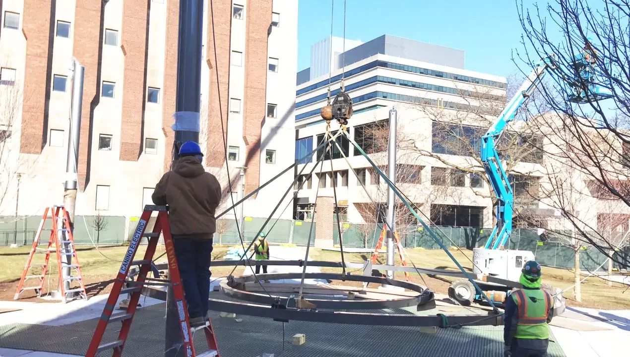 Construction workers use cranes and ladders to assemble a large metal structure on a lawn near multi-story office buildings.