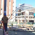Construction workers use cranes and ladders to assemble a large metal structure on a lawn near multi-story office buildings.