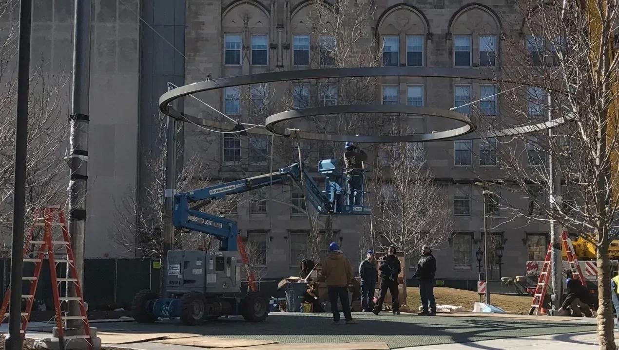Workers use a lift and ladders to install a large circular metal structure outdoors in front of a stone building with arched windows.