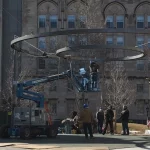 Workers use a lift and ladders to install a large circular metal structure outdoors in front of a stone building with arched windows.