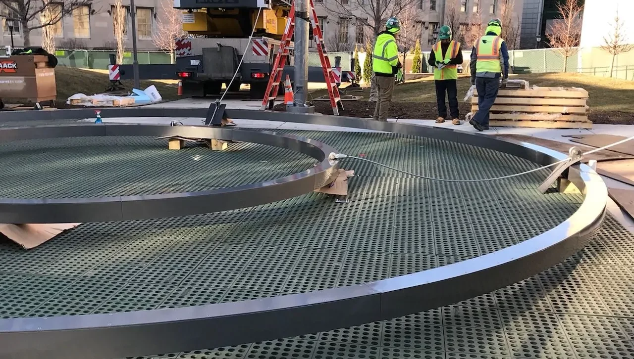 Large metal rings are positioned on a grated platform while construction workers in safety vests and helmets discuss nearby at an outdoor site with equipment and a crane in the background.