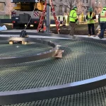 Large metal rings are positioned on a grated platform while construction workers in safety vests and helmets discuss nearby at an outdoor site with equipment and a crane in the background.