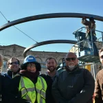 A group of six people pose in front of a large circular metal structure while a worker on a lift installs part of the structure under a clear blue sky.