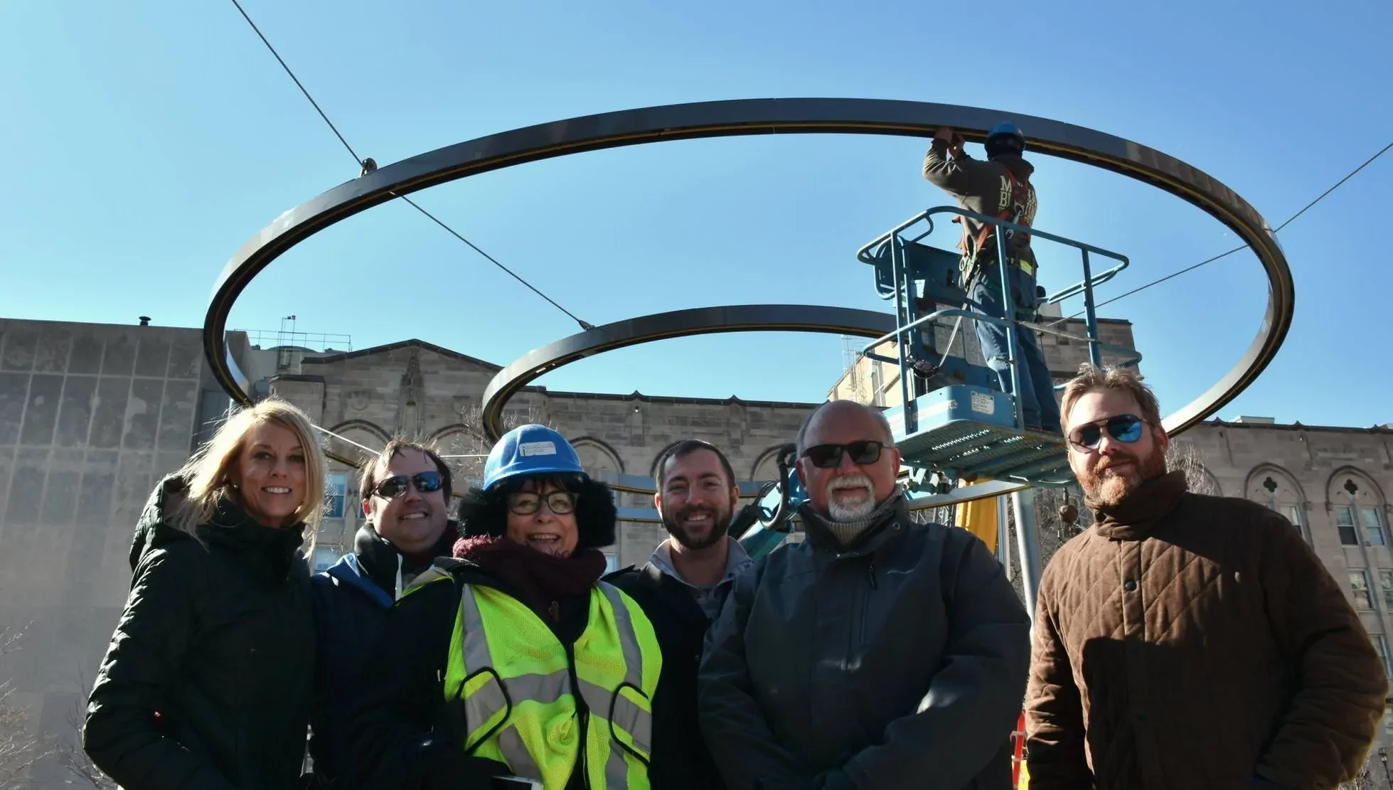 A group of six people pose in front of a large circular metal structure while a worker on a lift installs part of the structure under a clear blue sky.