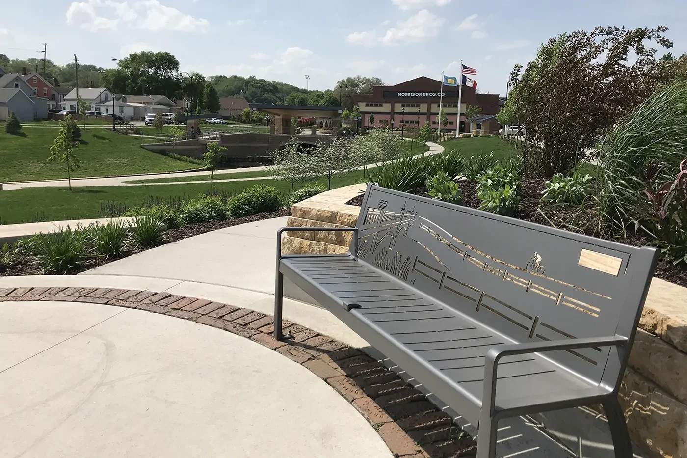 A metal bench with decorative cutouts sits on a curved sidewalk in a landscaped park with buildings and flags visible in the background.