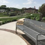 Un banc métallique avec des découpes décoratives se trouve à côté d'une allée courbe dans un parc paysager près d'un bâtiment scolaire et de mâts de drapeaux par une journée ensoleillée.