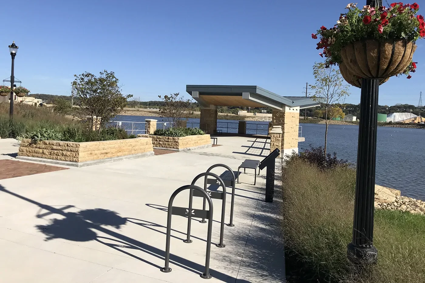 A paved riverside walkway with bike racks, benches, stone structures, a lamp post with hanging flowers, and water in the background on a sunny day.
