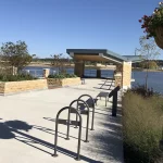Une promenade pavée le long de la rivière avec des porte-vélos, des bancs, une structure ombragée et un lampadaire avec un panier de fleurs suspendu au bord d'un plan d'eau calme lors d'une journée ensoleillée.