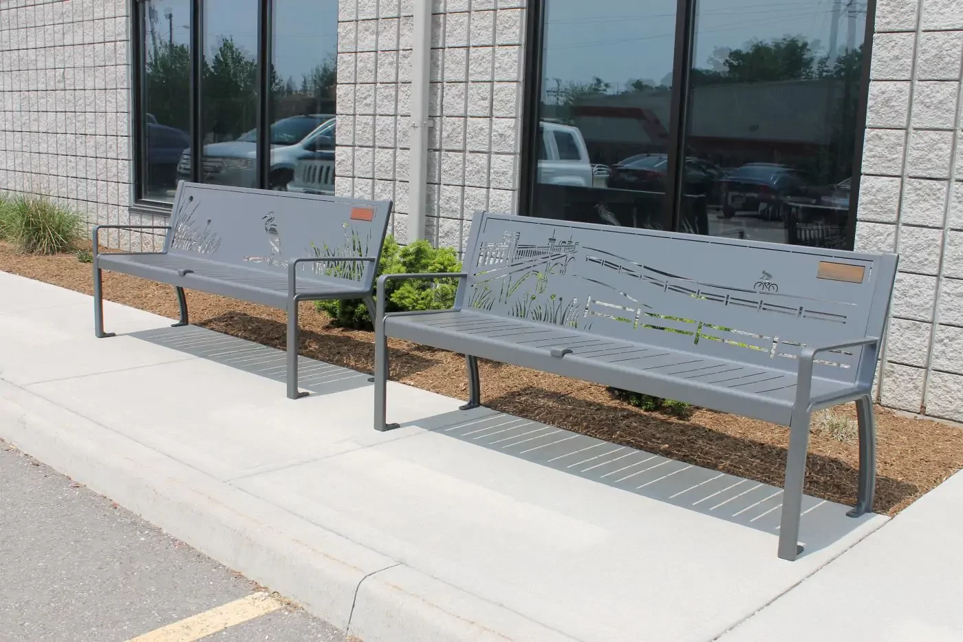 Two metal benches with cutout nature designs are placed on a sidewalk outside a building with large windows and light-colored brick walls.