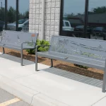 Two metal benches with cutout nature designs are placed on a sidewalk outside a building with large windows and light-colored brick walls.