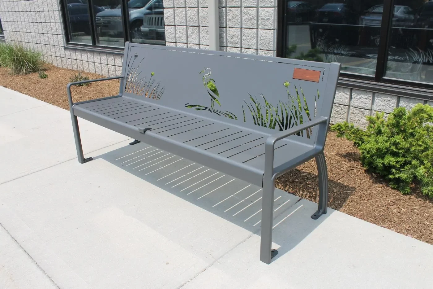 A gray metal bench with a backrest featuring cutout designs of a bird and tall grass, placed on a concrete sidewalk near a building and some shrubs.