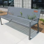 A gray metal bench with a backrest featuring cutout designs of a bird and tall grass, placed on a concrete sidewalk near a building and some shrubs.