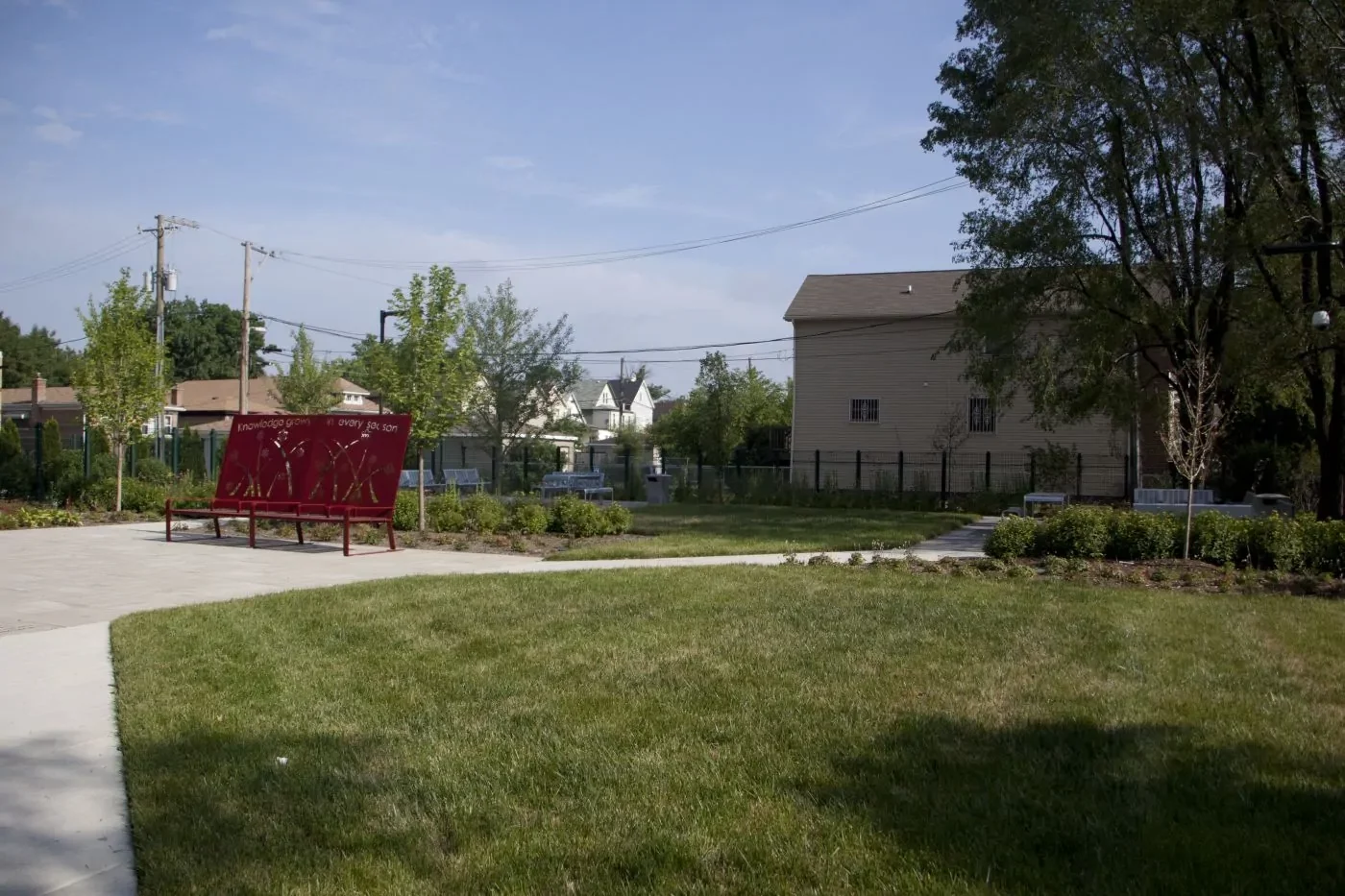 A small park with green grass, trees, a red bench, concrete walkways, and residential houses in the background under a clear sky.