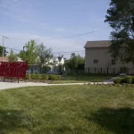 A small park with green grass, trees, a red bench, concrete walkways, and residential houses in the background under a clear sky.