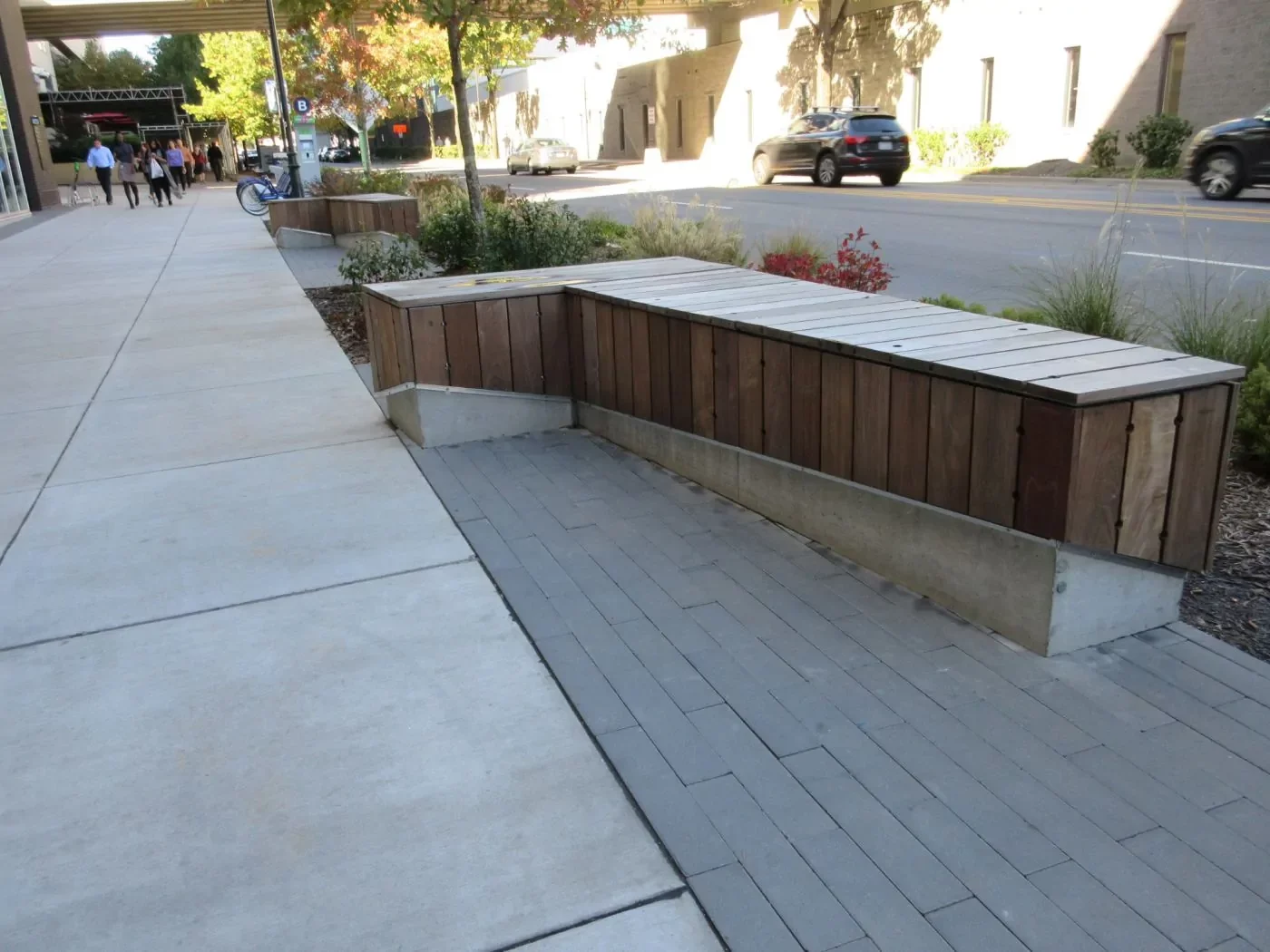 A wooden and concrete bench with planter boxes is situated along a sidewalk next to a street with some plants, trees, and parked cars visible.