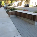 A wooden and concrete bench with planter boxes is situated along a sidewalk next to a street with some plants, trees, and parked cars visible.